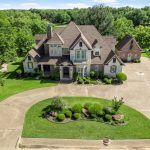 Aerial view of a two-story stone and brick home with a circular driveway, landscaped island, detached garage, and mature trees